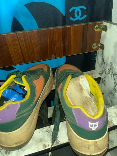 The scene depicts a pair of multi-colored athletic shoes resting on a dirty, light-colored surface with a mottled, marble-like pattern. The shoes are primarily dark green with orange panels, yellow ankle trim, and purple heel counters. The right shoe's heel features a white, stylized fox head logo. Dark green laces are present, with one untied and draped. The soles are thick, light tan, and appear dirty. The interior of the left shoe contains blue plastic material, while the right shoe's interior lining is visible and soiled. Above the shoes, a horizontal wooden panel with a brown grain is present, secured on the right by two brass-colored metal brackets. Behind and above this panel, a dark blue or black fabric is visible, displaying light blue graphics, including a partially obscured interlocking "CC" logo. Other dark, indistinct objects are partially visible beneath the shoes. This scene is located in Ilishan, Nigeria. No people are visible.