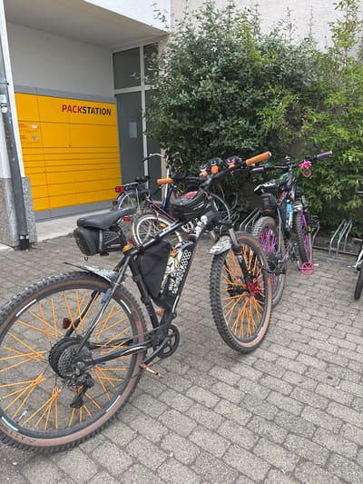 An outdoor shot shows a row of bicycles parked on a paved surface in front of a yellow "PACKSTATION" (package station) locker system. The lockers are arranged in horizontal rows and have individual compartments. To the right of the lockers is a dense green hedge. The bicycles appear to be mountain bikes, some with brightly colored spokes (orange and pink) and custom accessories. One bicycle in the foreground has a black frame with white graphics and a black saddlebag. Another bicycle has pink spokes and pedals. The pavement is made of grey interlocking bricks. The overall scene suggests a public area or residential complex where residents might use the package station and bicycle storage. The scene is located in Sindelfingen, Germany.