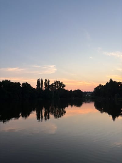 The image depicts a tranquil scene at dusk on a body of water, likely a lake or a wide river, bordered by a dense line of trees. The sky transitions from a pale blue at the top to soft hues of pink and orange near the horizon, indicating the setting sun. Wispy clouds are scattered across the sky, catching the last light of the day.
The water's surface is calm, reflecting the colors of the sky and the dark silhouettes of the trees along the shoreline. Several tall, slender trees stand out prominently in the center of the treeline, their shapes sharply defined against the fading light. On the far side of the water, beyond the immediate tree line, a few buildings with dark roofs are visible, suggesting a settlement or a rural area.
The overall atmosphere is one of peace and quiet, typical of the late evening in a natural setting. The location is identified as Rosnówko, Poland. There are no people or animals visible in the image. The reflection on the water is slightly rippled, creating a gentle distortion of the overhead scene.