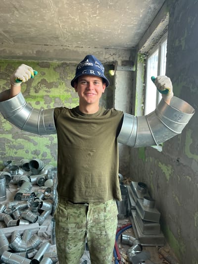 The image shows a young man, likely a construction worker, standing in a room under renovation. He is wearing a green t-shirt, camouflage pants, a blue bucket hat with "Rethymno" written on it, and white gloves. The man has two pieces of silver ductwork attached to his arms, resembling pipe extensions, and is holding a small green object in each gloved hand. He is smiling and looking at the camera.
The room appears to be unfinished, with exposed concrete ceilings and walls that are partially painted with a distressed green color. The floor is concrete and littered with various pieces of silver ductwork, including elbows and straight sections, as well as some plumbing pipes. In the background, there is a window and more ductwork stacked in the corner. The overall impression is a construction site or a storage area for building materials. The location is specified as Аснежыцкі сельскі Савет, Belarus.