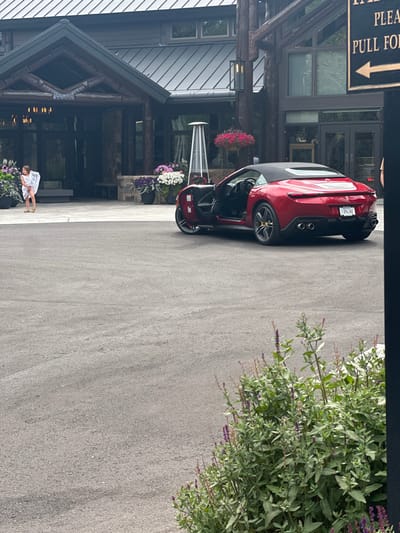 A mid-range outdoor photograph depicts the paved entrance area of The Little Nell Hotel in Aspen, Colorado, United States. A dark red Ferrari Portofino convertible is centrally parked, facing away from the viewer, with its driver's door open and convertible top closed. The vehicle displays Colorado license plate T-89436E and features yellow brake calipers. To the left, near the hotel's timber and stone entrance, a young female child in a light-colored garment stands, facing left. The background features the multi-story hotel building, adorned with various potted flowers. A silver patio heater is positioned near the vehicle. A partial sign on the far right reads "PLEASE PULL FOR," indicating valet service. The immediate foreground, lower right, contains a bush with green leaves and purple flowers. The scene is illuminated by natural, diffused light.