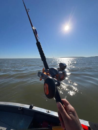 A first-person perspective shot captures a fishing rod with an orange reel and a purple lure against a bright blue sky. The sun shines intensely, casting a glare on the water. The scene appears to be from a boat on a body of water, possibly a lake or river, with a distant shoreline visible. The foreground shows the edge of the boat and a hand holding the fishing rod. The image does not provide enough visual information to definitively place it in Novopichugovo, Russia.