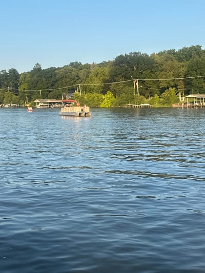 The image captures a serene summer day on a lake in Algonquin, United States of America. The foreground is dominated by the tranquil blue water of the lake, with gentle ripples reflecting the clear sky. In the mid-ground, a pontoon boat with several people on board cruises across the water, its canopy providing shade. Nearby, a red buoy marks the waterway. The background reveals a lush, tree-lined shore with houses and docks, suggesting a residential area bordering the lake. The sky is a clear, bright blue, typical of a sunny day. The overall atmosphere is one of peaceful leisure and recreation.