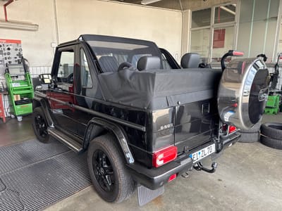 A black Mercedes-Benz G 500 convertible is parked indoors, likely in a car repair shop or garage. The vehicle has its soft top up, covering the rear passenger area and trunk. The spare tire is mounted on the rear of the vehicle, with a visible license plate that reads "EI ZL 70". Next to the car, a stack of tires is visible. In the background, a green Bosch diagnostic machine is present, along with a wall-mounted chart detailing car parts. The floor is a concrete surface with a metal grate in front of the vehicle. The environment suggests a service or maintenance setting.