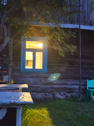 A rustic wooden building with a blue-framed window illuminated from within is the central focus of the image. A tree with foliage partially obscures the window and the building's facade. In the foreground, the corner of a light-colored table with a mottled surface is visible, reflecting the interior light. To the right of the building, a turquoise bench is situated in front of a patch of green grass, which transitions into darker, rougher terrain. The overall impression is that of a rural setting at dusk.