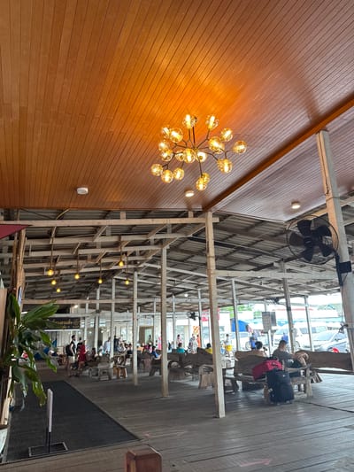 This is a wide-angle, eye-level shot of an open-air building with a wooden slatted ceiling and wooden pillars supporting a corrugated metal roof. The space appears to be a waiting area or a casual restaurant, possibly at a pier or ferry terminal in Ban Phe, Thailand. A large, modern chandelier with multiple glass globe lights hangs from the wooden ceiling. Several smaller, yellow pendant lights on ropes are suspended from the corrugated roof structure. A black industrial fan is mounted on one of the white wooden pillars.

The floor is made of worn wooden planks. Several wooden benches, some with rough, natural wood slabs as backs and seats, are arranged throughout the space. People are scattered throughout the building, some sitting on the benches, others standing and conversing. In the foreground left, a large potted plant with broad green leaves is visible.

To the right, a couple sits on a bench with luggage, including a black wheeled suitcase and a red duffel bag. In the background, there are glimpses of vehicles, possibly cars and vans, suggesting proximity to a road or transportation hub. Signs with Thai script are visible on some of the walls. The overall atmosphere is relaxed and casual, with natural light filtering into the space, complemented by the artificial lighting.