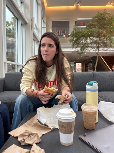 The image depicts the interior of a modern institutional building, likely a college or university common area or student center in Wellesley, United States.

A woman with long, dark brown hair and light skin sits on a dark gray ribbed sofa in the foreground, facing slightly to the right of the frame. She wears a light yellow knitted sweater with red lettering and blue jeans. Her left hand holds a partially eaten sandwich, while her right hand holds a piece of crumpled brown paper. Her gaze is directed upwards and to the left, with her mouth slightly open, suggesting she is chewing or about to take a bite. Numerous rings adorn her fingers, and bracelets are on her wrist.

On the dark rectangular table in front of her are several items: a white paper coffee cup with a brown sleeve, a clear plastic cup containing an iced beverage with layered liquids, a tall light yellow water bottle with a blue and white cap, and various pieces of crumpled brown and white paper food wrappers. A silver laptop is partially visible on the right side of the table.

The background reveals a spacious, brightly lit interior with tall, floor-to-ceiling windows on the left, showing a blurry outdoor view. The building features high ceilings with recessed lighting and a multi-level design with a visible upper-level balcony. An indoor tree with light-colored bark and green leaves stands prominently in the mid-ground. A modern staircase with dark steps ascends on the right, and a red "EXIT" sign is visible above it. Blurry figures are present on the upper-level balcony and near the staircase, suggesting other occupants of the space.