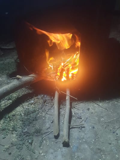 A nighttime outdoor scene depicts a rustic, metal fire pit containing a vibrant, roaring fire. The flames are intensely bright, casting an orange and yellow glow on the surrounding area. Two long, thin wooden sticks are placed in front of the fire pit, angled towards the flames. To the left of the fire pit, a thick, gnarled tree branch lies horizontally on the ground. The ground itself is covered in a mixture of dirt, small stones, and dried leaves. The overall impression is one of a simple, possibly rural, setting where a fire is being actively maintained for warmth or cooking.