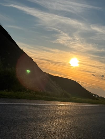 This image captures a sunset scene with a prominent lens flare. A dark, sloped hill covered in vegetation dominates the left side of the frame, obscuring much of the landscape. The sun is setting on the right, casting a warm orange glow across the sky and creating streaks of light in the clouds. A bright, green circular lens flare is visible on the hillside. In the foreground, a paved road with a white line runs horizontally, reflecting the sunset's light. The environment appears to be rural or semi-rural, with hints of distant vehicles on the right side of the image. The location is identified as Таймурзино, Russia.