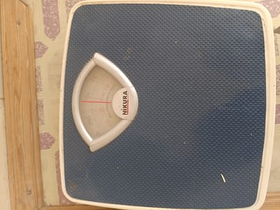 This is a top-down, close-up photograph of a blue analog bathroom scale situated on a patterned floor. The scale has a white bezel and a central dial with markings for both pounds (lb) and kilograms (kg). The dial shows weight readings up to 260 lb and 130 kg. The brand name "NIKURA JAPAN" is visible on the dial. The scale is placed next to a wooden plank, and the floor appears to be tiled with a repeating geometric pattern in shades of beige and purple. There are some small specks of dirt or debris on the surface of the scale. The lighting suggests it is daytime. The overall impression is of a functional, albeit slightly worn, household object in a simple setting.