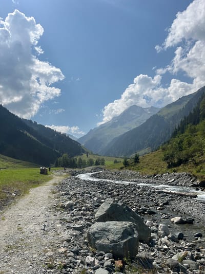 The image displays an alpine valley in Reitlehen, Austria, under a bright blue sky with scattered cumulus clouds. A wide, rocky riverbed, containing a meandering stream, extends from the foreground into the distant background on the right side of the frame. Two large, light gray boulders are situated prominently in the immediate foreground on the riverbed. Parallel to the riverbed, on the left, a light-colored gravel path traverses a section of green grass. In the middle ground, three to four indistinct human figures are observed walking away from the viewer on this path. Adjacent to the path, near these figures, is a stack of dark brown, possibly wooden, material. The valley is flanked by steep, heavily forested mountainsides that converge in the hazy distance. Direct sunlight illuminates the scene, creating distinct shadows.