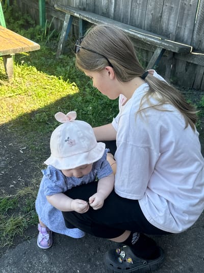 In Novoyeniseyka, Russia, a young girl with blonde hair tied in a ponytail, wearing a white t-shirt and black pants, crouches to interact with a baby. The baby, wearing a light pink sun hat with a flower detail and a blue dress, is also crouched down. The baby is holding something small in their hands. The scene is outdoors, with a wooden fence and vegetation visible in the background. A wooden picnic table is partially visible on the left. The ground is a mixture of dirt and grass. The lighting suggests it is daytime, with shadows indicating sunlight.