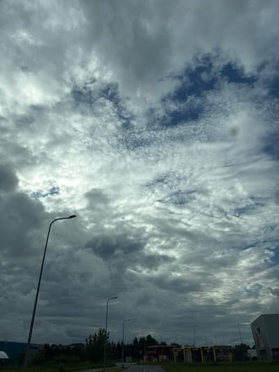 The image depicts a sky filled with a dramatic mix of dark and light gray clouds, with patches of blue sky peeking through. The clouds are textured and varied, suggesting a dynamic weather system. In the foreground, a streetlamp with a dark, curved pole stands on the left side of the frame, silhouetted against the sky. Further back, more streetlamps are visible along a road that disappears into the distance. On the right side of the image, a modern, angular building with a light-colored facade is partially visible. The lower part of the image shows a stretch of green grass and trees, with what appear to be structures and possibly toll booths or kiosks lining the road. The scene is located in the town of Peetri alevik, Estonia, under a cloudy sky.