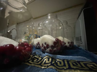 In a dimly lit kitchen, possibly located in Akhtyrskiy, Russia, a large glass jar filled with bright red cherries and white sugar sits prominently in the foreground. The jar, partially obscured by a deep red pile of cherries, has a distinctive patterned blue and gold cloth draped in front of it, suggesting a decorative or functional purpose, perhaps to absorb any potential leaks or for aesthetic appeal.
Behind this primary jar, three more identical glass jars are arranged in a row. Each of these jars contains a layering of red cherries and white sugar, indicating they are in the process of preservation or preparation for a recipe, likely involving fruit preservation. The jars have a threaded opening, suggesting they will be sealed with lids.
The background reveals a tiled white backsplash, typical of a kitchen environment. To the right, a portion of a silver appliance, possibly a microwave or refrigerator, is visible, its dark surface contrasting with the lighter kitchen elements. The overall impression is one of domestic food preparation, with an emphasis on preserving fresh produce. The lighting is low, creating a slightly shadowed and intimate atmosphere. No people are visible in the frame.