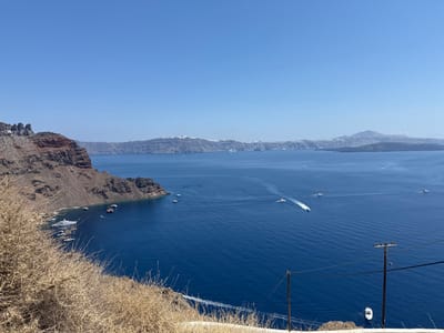 The image depicts a panoramic view of the Santorini caldera, specifically located within the Ia Municipal Unit, Greece. The scene features a deep blue Aegean Sea under a clear, cloudless sky. To the left, a prominent dark and reddish volcanic cliff face, part of the caldera wall, descends steeply to the water's edge. Numerous white boats and yachts of varying sizes are anchored or moored near the base of this cliff. Across the expansive blue water, several smaller motorboats are observed in motion, leaving distinct white wakes behind them, indicating travel. In the distance, the outlines of volcanic islands, characterized by their dark, barren appearance and some snow-capped peaks, are visible on the horizon, defining the caldera's rim. In the lower right foreground, two dark wooden utility poles with associated power lines are present. The immediate foreground, particularly on the left, consists of dry, light brown, bushy vegetation. No discernible human figures are detailed.