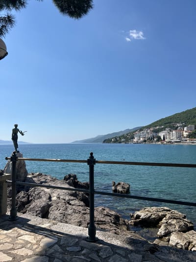 This media file presents a scenic coastal landscape, likely captured on a bright, clear day in Grad Opatija, Croatia. The image is oriented to show a vast expanse of clear blue sky at the top, with a small, wispy white cloud in the upper left quadrant.
The horizon line spans diagonally across the mid-ground, separating the sky from the land and sea. On the left, a lush, green hillside rises, densely covered with trees and a collection of white and light-colored buildings, characteristic of a vibrant coastal town. This urban area stretches along the shoreline, indicating a settlement nestled against the verdant slopes.
To the right of the horizon, the calm, blue-green waters of the sea extend into the distance, reflecting the clear sky. In the immediate foreground, a dark metal railing runs horizontally, providing a vantage point over the water. Prominently featured below this railing, and partially silhouetted against the bright sky and sea, is a bronze statue known as the "Maiden with the Seagull" (Djevojka s galebom). The elegant female figure stands on a pedestal, holding out her arm with a bird (a seagull) perched or taking flight from her hand.
To the right of the statue and railing, the rugged coastline is visible, composed of large, dark rocks, some partially submerged in the transparent water. A patterned stone path or wall, casting sharp shadows, occupies the lower right corner of the frame. The strong, direct sunlight suggests it is either morning or early afternoon. No people or visible text are present in the image, conveying a sense of tranquility and natural beauty.