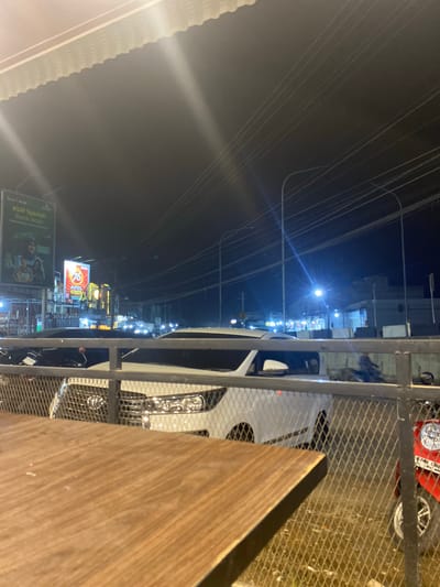 The image depicts a nighttime scene in Tanjongkapai, Indonesia. A wooden table is visible in the foreground, along with a metal structure resembling a fence or railing. A white car is parked on the street. In the background, there is a street with various signs, including one for "KUR Syariah Bank Aceh" and another for "76". Streetlights illuminate the scene, and power lines stretch across the dark sky. A red object with a headlight is visible below the fence, possibly a scooter.