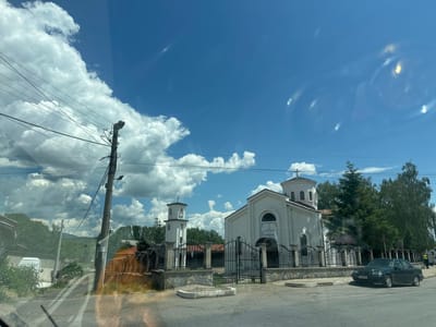 The image shows a street view in the city of Nevestino, Bulgaria. On the right side of the street, a white church with a bell tower stands behind a black metal fence and a stone wall. The church has a dome and a cross on top. In front of the church, a dark green Mercedes-Benz sedan is parked on the side of the road. Trees are visible behind the church and to the right of the scene. On the left side of the street, a utility pole with wires is present, and a white van is parked in front of a building. The sky is blue with scattered white clouds.