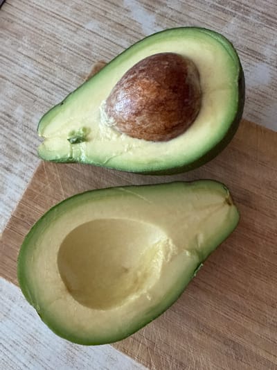 The media file displays a top-down view of two halves of an avocado on a wooden surface. The avocado on the left is whole and still contains its large brown pit, while the avocado on the right has been halved, revealing the smooth, pale green flesh and the empty cavity where the pit would have been. The avocados have a dark green skin with some lighter green undertones. The background is a light-colored wooden surface with a subtle wood grain pattern. To the right of the avocados, a distinct wooden cutting board with a more pronounced grain and a slightly darker hue is visible, suggesting the avocados have been prepared or are about to be prepared. The lighting appears to be natural, casting soft shadows. No people are present in the image, and the focus is solely on the avocados and their immediate surroundings. The scene is simple and domestic, likely a kitchen setting. There is no visible text in the image. The location context provided is Kavkazkaya, Russia.