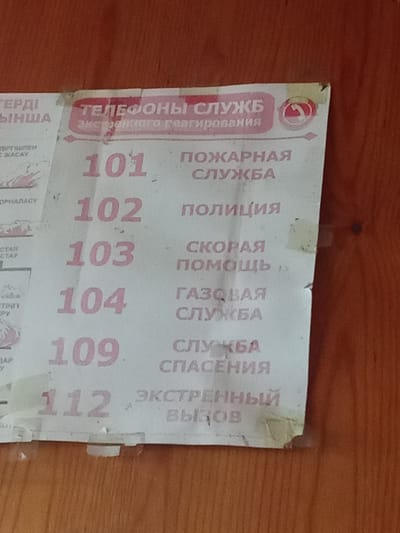 The image shows a piece of paper with emergency telephone numbers listed in Russian and what appears to be Kazakh. The paper is attached to a wooden surface, possibly a door or wall, with tape. The paper is worn and slightly torn in places.

The listed numbers and their corresponding services are:
- 101: Пожарная служба (Fire service)
- 102: Полиция (Police)
- 103: Скорая помощь (Ambulance)
- 104: Газовая служба (Gas service)
- 109: Служба спасения (Rescue service)
- 112: Экстренный вызов (Emergency call)

There are also some lines of text in what appears to be Kazakh script to the left of the numbers, but they are partially obscured and difficult to read. The overall impression is one of a practical informational sign posted in a public or residential area. The lighting suggests an indoor setting during daylight.