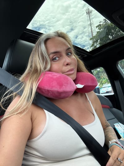A woman with blonde hair, streaked with grey, is seated in a car. She is wearing a white tank top and a pink neck pillow. A black seatbelt is fastened across her chest. Her left arm is resting on the seatbelt, and her hand is visible with rings on her fingers and a teal bracelet. A package of something is visible on the car seat to her right. The car has a sunroof open, revealing a cloudy sky with a tall communication tower and green trees visible. The scene appears to be in Tambon Pak Phraek, Thailand, suggested by the presence of the communication tower and lush vegetation typical of the region.