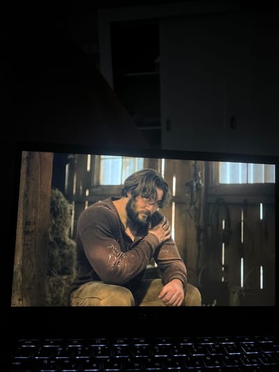 The image captures a scene viewed on a screen within a dimly lit room, with a backlit keyboard visible to the right. The screen displays a man in what appears to be a rustic or historical setting. The man has long, dark hair and a full beard, wearing a dark brown, long-sleeved shirt with some light speckles or debris on it, and what looks like light-colored trousers. He is hunched over, seemingly in distress or deep contemplation, with one hand gripping his other arm or chest. The background behind him features wooden planks, suggesting an old barn or cabin, with natural light filtering through horizontal slats, possibly from a window. A pile of hay or straw is visible beneath him.

The environment outside the screen is dark, emphasizing the glow of the display and the keyboard. The keyboard, positioned vertically along the right edge of the frame, features a standard QWERTY layout with additional Cyrillic characters, illuminated by backlighting. Visible keys include "esc", "tab", "caps lock", "shift", "ctrl", "alt", "enter", and various letters and numbers in both Latin and Cyrillic scripts (e.g., 'й', 'ц', 'у', 'к', 'е', 'н', 'г', 'ш', 'щ', 'з', 'х', 'ъ', 'ф', 'ы', 'в', 'а', 'п', 'р', 'о', 'л', 'д', 'ж', 'э', 'я', 'ч', 'с', 'м', 'и', 'т', 'ь', 'б', 'ю'). The overall impression is someone watching a film or show on a laptop or tablet at night, in Voysil, Bulgaria.