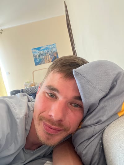The image shows a selfie taken by a man lying in bed. He is partially covered by a gray blanket and a pillow. The man has light brown hair, green eyes, and stubble on his face. He is wearing a gray t-shirt. The background appears to be a bedroom with a light-colored wall and a picture of a cityscape hanging on it. There is also a baby crib in the background, suggesting the man might be a parent. The lighting suggests it could be daytime. The man's expression is relaxed, possibly indicating he is waking up or lounging in bed. No visible text is present in the image. The location context is Rumia, Poland.