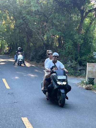 In Kubu, Indonesia, on a paved road surrounded by lush green foliage and trees, three people are riding a scooter. A man, wearing a white t-shirt, sunglasses, and a white head covering, is driving the scooter. A woman, with sunglasses and wearing a white top, is riding behind him. A child is seated in front of the man. Another scooter, a white one, is visible in the background with two individuals riding it. The road has yellow markings. A sign with the word "alani" is visible on the right side of the frame.