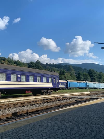 This image depicts the Yasinia train station in Yasinia, Ukraine, under a clear blue sky with scattered white cumulus clouds. In the foreground, a grey paver-stone railway platform extends from the lower right, casting a shadow. Adjacent to the platform are multiple parallel railway tracks laid on gravel ballast with concrete sleepers.

A stationary dark blue passenger train carriage with a cream-colored horizontal stripe occupies the tracks closer to the platform. This carriage features multiple windows with light-colored curtains and displays the Ukrainian Railways (UZ) logo along with the numbers "043" and "22350." Further along the tracks, behind the passenger car, a series of light blue freight train cars, including several open-top or hopper-style cars, are visible.

The background consists of a mountainous landscape characterized by densely forested green hills. Utility poles and power lines are visible across the mid-ground, running parallel to the railway lines. No individuals are present in the scene. Sunlight illuminates the entire scene, casting distinct shadows on the platform and tracks.