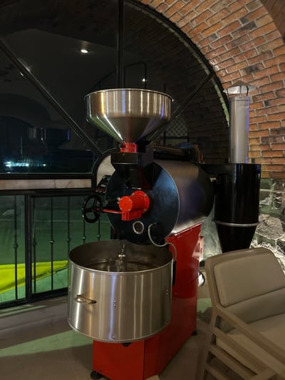 The image shows a coffee roasting machine inside a building in Santa Ana Yancuitlalpan (Chilhuacán), Mexico. The machine is primarily black and silver, with red components. It has a cylindrical roasting drum on the right and a conical hopper on the left. A ventilation system with a silver and black chimney extends upwards. The machine is positioned near a large arched window with metal framing. Behind the machine is a brick wall and part of a chair with white cushions. The floor is light-colored. The environment appears to be indoors, likely a cafe or roastery.