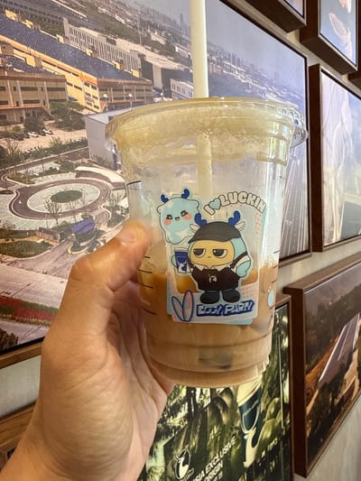 A hand is holding a disposable plastic cup filled with a light brown beverage and a white straw. A sticker with a cartoon character is affixed to the cup. The background is a series of framed pictures. One picture appears to depict an aerial view of a city with modern architecture and a circular traffic intersection. The scene is likely located in Changping District, China, based on the cityscape depicted in the background picture.