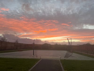 A serene twilight view captures a tennis court with nets, set against a sky ablaze with vibrant hues of orange, pink, and grey clouds. The court's grey surface reflects the fading light, and a dark pathway leads into the scene. A brick wall, trees, and green grass border the court, adding to the peaceful, almost surreal, ambiance of dusk.