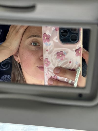 A self-portrait captured via reflection in a car's vanity mirror in Karagay, Russia. The image features the upper face of an individual, presenting a greenish-blue left eye, a defined eyebrow, and partial views of the nose, mouth, and a freckled cheek. The subject's right hand is positioned on the forehead. A small gold-colored hoop earring is visible in the left ear. A light pink smartphone, featuring a multi-lens camera array and encased in a floral-patterned "Casetify" case, is held vertically by the subject's left hand, obscuring a portion of the face. This hand displays a white braided ring on the middle finger and a second ring on the ring finger. The reflected background behind the subject's head consists of a dark fabric with a white circular pattern, consistent with vehicle interior upholstery. The grey frame of the sun visor mirror borders the reflective surface.