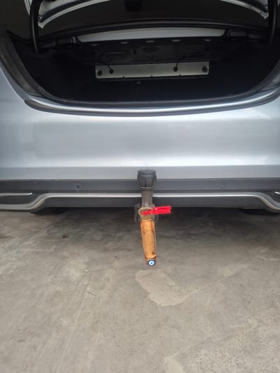 A rear view of a silver vehicle is shown, with the trunk open. The vehicle has a tow hitch attached. A decorative, wood-carved object with a blue evil eye charm is hanging from the tow hitch. A red clothespin is attached to the tow hitch. The scene appears to be outdoors on a concrete surface, possibly a driveway or parking lot, in Hochspeyer, Germany.