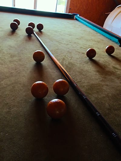 The image depicts an indoor scene in Терновский округ, Ukraine, featuring a billiard table. The table is covered with an olive-green felt cloth, exhibiting visible texture and minor surface irregularities. A dark green rail forms the upper edge of the table. Twelve wooden or amber-colored billiard balls are distributed across the felt surface; one ball in the mid-left section has a distinct horizontal groove. A billiard cue stick lies diagonally on the table, extending from the upper-center towards the lower-right. The cue stick transitions from a lighter wood shaft to a darker, possibly stained or wrapped, butt end adorned with etched rings. In the upper left background, a window is visible, providing ambient light. To the upper right, a reddish-brown wooden wall is present, against which a large, white, curved object is partially visible on the far right. The illumination, originating from the upper left, casts shadows from the balls and the cue stick onto the table's felt surface, most notably in the lower right portion of the image. No individuals are present.