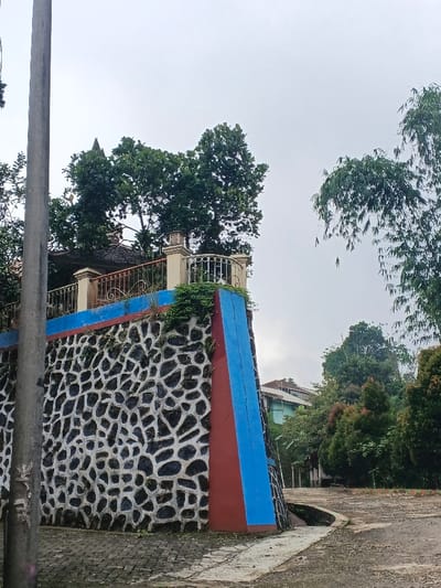 The image depicts a street scene in Makilala, Philippines, characterized by a prominent retaining wall. The wall, forming a corner, features a lower section composed of irregularly shaped dark stones set in white mortar. Above this, the wall is painted with horizontal bands of red and blue. The top of the wall supports a structure with light-colored pillars and a white metal balustrade, indicative of an elevated patio or balcony, from which dense green foliage of trees is visible. A grey utility pole occupies the left foreground. The ground level transitions from a paved, possibly cobblestone-patterned surface in the immediate foreground to a dirt or gravel path extending to the right. A narrow concrete gutter runs along the base of the wall. To the right, various trees and shrubs with green and reddish foliage are present. In the background, partially obscured by vegetation, are residential structures with light-colored walls and sloped roofs. The sky is overcast and light grey, suggesting a cloudy day. No individuals or specific actions are visible.