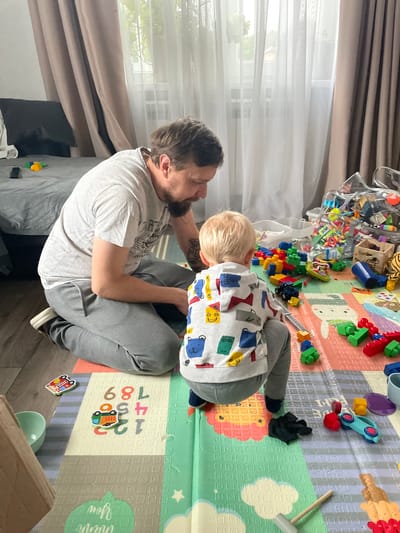 The image shows a father and his young son playing with toys on a playmat in a room. The father, with a beard and tattoos on his arm, is wearing a grey t-shirt and grey sweatpants. The son, with blonde hair, is wearing a white t-shirt with cartoon cars on it and grey sweatpants. They are both sitting on a colorful playmat covered with various toys, including building blocks, a toy car, and toy figures. The playmat has numbers and cloud shapes printed on it.

In the background, there are sheer white curtains and brown drapes, suggesting the room has a window. The overall scene conveys a sense of playful interaction and familial bonding. The lighting appears to be natural, possibly from a window, and there are no strong indicators of the time of day or weather outside. The location context provided is Субботино, Russia. There is no visible text in the image that provides additional context or information.