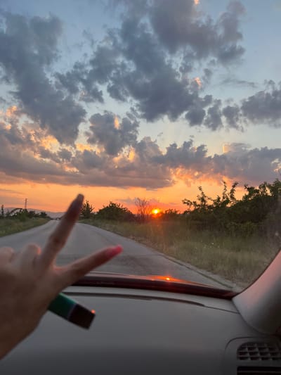 The media file captures a scenic sunset viewed from inside a car, possibly during a road trip. The sky is filled with dramatic clouds, painted in hues of orange, pink, and gray as the sun dips below the horizon. The setting sun casts a warm glow on the landscape, which appears to be rural, with trees and vegetation lining a winding road. A hand, adorned with bright pink fingernails, is visible in the foreground, making a peace sign. A vape pen, colored green and black, is held between two fingers of the same hand. The interior of the car, specifically the dashboard and windshield frame, are also visible, indicating the viewpoint. The time of day is clearly sunset, given the vibrant colors of the sky and the low angle of the sun. The weather appears clear, with the clouds adding to the visual appeal of the sunset. While no specific text is visible, the overall impression is one of leisure and enjoyment of a beautiful natural spectacle. The context provided places this scene in Velichkovo, Bulgaria.