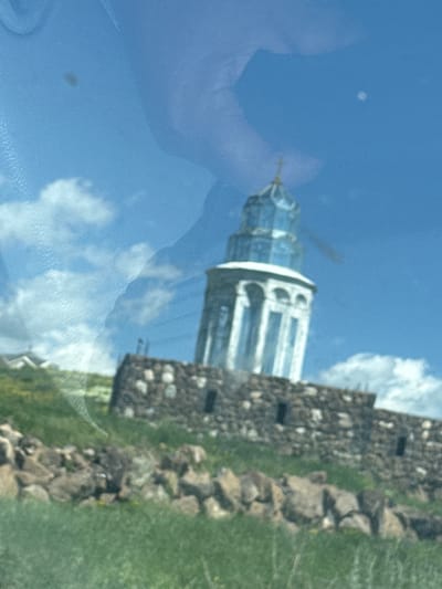 The image shows a lighthouse on a rocky hill with a bright blue sky and some clouds. There are reflections on the surface, obscuring parts of the image. The lighthouse has a white base with arched windows and a blue, glass-like top. The hill is covered with grass and scattered rocks. The image appears to be taken from a moving vehicle. This is likely the lighthouse in Artashavan, Armenia, situated on a hill overlooking a body of water, possibly Lake Sevan. The reflection in the foreground is unclear, but it could be a person or the interior of a vehicle. There are no clear interactions or actions visible due to the reflections and blurriness.