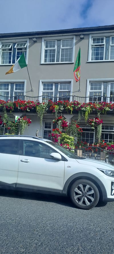 The image shows a white car parked on a street in front of a building adorned with abundant red and white flowers. Two flags are visible hanging from the building's facade. The flag on the lower left is the Irish tricolor, featuring green, white, and orange bands. Above it, another flag, partially visible, displays red, yellow, and green colors, possibly representing a different national or regional symbol. The building has multiple windows with white frames, and some of them are open. The architecture suggests an older European style, and the abundant floral decorations create a picturesque and vibrant scene. The car, a modern SUV, is parked close to the building, its side and wheel clearly visible. The ground is paved with asphalt. The overall impression is a bright, sunny day, likely late morning or afternoon, contributing to the cheerful atmosphere created by the flowers and flags. The location context of Westport, Ireland, aligns with the presence of the Irish flag and the architectural style.