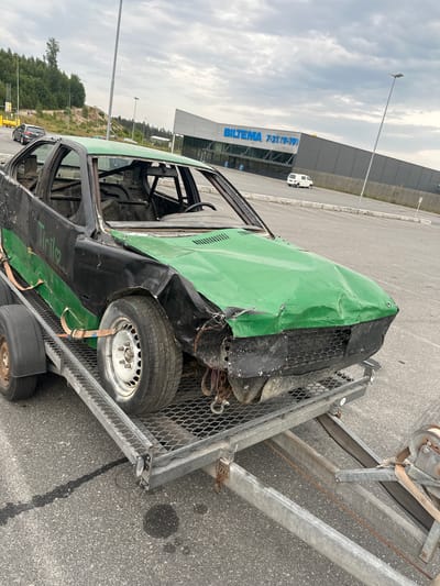 An exterior, medium-shot, eye-level, daytime, overcast view shows a heavily damaged black and green car on a gray metal trailer. The car's roof and windows are removed. The hood is crumpled and painted bright green, and the front bumper is missing, exposing wires and mechanical components. The car has a roll cage and a spare tire visible inside. The words "Tiril" are written in black marker on the driver's side door, with a heart symbol next to it. The trailer is secured with orange straps to the car's wheels. The trailer is parked on an asphalt surface, with a faint black fluid stain visible on the ground. In the background, a large, gray building with blue lettering that reads "BILBEMA 7-21 (9-19)" is visible, suggesting a retail store. A white van is parked in the store's lot. The location is identified as Slitu, Norway.
