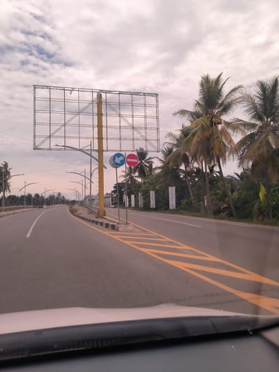 The image captures a road scene from the perspective of a vehicle's interior, likely driving in Meunasahpulo, Indonesia. The paved asphalt road curves gently to the left, leading onto what appears to be a bridge or overpass, lined with modern streetlights. White solid and broken lines mark the lanes, while prominent yellow diagonal hatching is visible on the right side of the road, indicating a specific traffic zone.
On the right side of the road, a large, empty metal billboard structure with a prominent yellow supporting pole stands tall. Below it, two circular traffic signs are mounted: a blue sign with a white arrow pointing left, typically indicating a mandatory turn or lane, and a red sign with a white horizontal bar, signifying "no entry" or "do not enter."
Beyond the road and structures, lush tropical vegetation is visible, dominated by numerous tall palm trees, some bearing visible coconuts. Smaller white banners or signs are interspersed within the foliage on the right roadside. The sky is overcast with scattered white and grey clouds, suggesting a cloudy day or possibly late afternoon/early morning due to the diffused lighting. The lower portion of the image shows the top of a car's dashboard and windshield wipers. There is no discernible text on any of the signs or banners, beyond the universal traffic symbols.