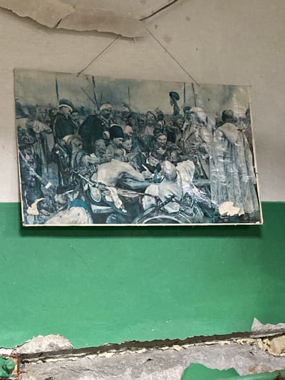 The image depicts a framed, aged, and slightly damaged painting hanging on a wall. The painting itself is a detailed depiction of a historical or military scene, featuring a large gathering of men in traditional attire, many with beards and wearing hats. There are visible elements of swords, shields, and spears within the painting. The painting is suspended by thin wires from the ceiling. The wall behind the painting is a light, possibly white or off-white color, and shows signs of peeling paint and water damage at the top. Below the painting, the wall is painted a vibrant green. The bottom edge of the image reveals a rough, concrete-like surface with more peeling paint and some visible fixtures, suggesting an interior space that may be undergoing renovation or is in a state of disrepair. This scene appears to be located in the city of Ukrainka, Ukraine, possibly within an older building or a public space like a community center or a historical site.