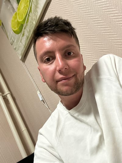 A man is looking down at the camera. He is wearing a white sweatshirt. Part of a lime-themed picture is visible in the bottom left corner. The location is a room with beige wallpaper and white pipes. There is no evidence to suggest this image was taken in Vorotynsk, Russia.