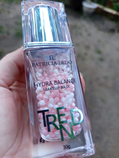 A person's hand holds a clear glass bottle filled with pink and white beads. The bottle has a silver cap and is labeled "PL PATRICIA LEDO HYDRA BALANCE MAKEUP BASE TREND 30g". The background is blurred and appears to be outdoors, with dirt and some greenery visible, suggesting a natural or garden setting. The lighting indicates it is daytime. The overall mood is neutral, focusing on the product.
