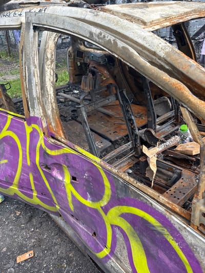 The image shows a heavily damaged and burnt-out car in Kokkorevo, Russia. The car's interior is gutted, with exposed metal frames of the seats and evidence of fire damage throughout. Graffiti in purple and yellow spray paint covers the exterior of the driver's side door. Debris, including charred material and a plastic bottle, is scattered around the vehicle. The surrounding environment appears to be an outdoor area with some greenery visible in the background and a brick structure. The scene suggests the car was abandoned or destroyed by fire.