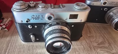 This image features a vintage FED 3 camera placed on a wooden surface, possibly a table or shelf. The camera is silver and black with a metallic lens. Several AAA batteries, some red and others white with red lettering, are scattered to the left of the camera. The batteries appear to be a mix of brands, including "Smartbuy" and "365 AAA HEAVY DUTY".
The camera itself has several dials and markings. Visible text on the camera includes "FED 3" in a stylized font, and Cyrillic lettering which appears to be part of the lens's specifications, such as "ИНДУСТАР" and numerical aperture and focal length markings like "2.8/52", "8 11 16", and "4 5 7 10 20". There are also numbers on a dial on the top of the camera, including "B", "250", "500", and "30", which likely represent shutter speed settings.
The overall setting is indoors, with a blurred red background suggesting fabric or another object behind the camera. The lighting is natural or soft artificial light, creating some reflections on the metallic surfaces of the camera. There are no people visible in the image. The scene appears to be a still life arrangement, possibly showcasing photographic equipment or an older collection. The presence of batteries suggests the camera might be in use or prepared for use. The location context is Gostagayevskaya, Russia, but there are no specific visual cues within the image to confirm this.