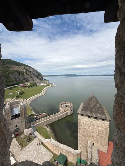 The image shows a view from a stone structure, looking out over a fortress situated on the edge of a large body of water, likely the Danube River, near Golubac Fortress in Golubac Municipality, Serbia. In the foreground, the rough texture of stone walls frames the scene.

Within the fortress complex, several stone towers and walls are visible. One prominent tower in the right-middle ground has a steep, dark, multi-faceted roof. To its left, a circular tower with battlements extends into the water. A stone walkway connects parts of the fortress. Moored in the water near the circular tower is a long, narrow boat with a wooden deck.

Beyond the fortress, a green, grassy area slopes upwards towards a rugged, tree-covered mountain. A paved road runs along the base of the mountain, with some buildings visible in the distance. The water stretches out to the horizon, meeting a sky filled with scattered white clouds.

Several individuals are present within the fortress grounds. Two figures in dark clothing walk on a paved area in the lower-middle left of the frame. Other figures are sparsely distributed throughout the visible courtyards and pathways. Two green tents are set up in the lower right portion of the image.