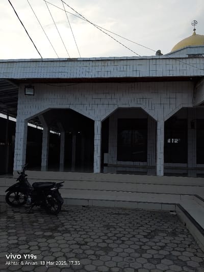 The image shows a view of the exterior of a mosque in Kecamatan Gabuswetan, Indonesia, with coordinates of -6.44 latitude and 108.028 longitude. The mosque is constructed of white tiled walls and has several arched doorways. The doorways are shaded and dark, leading to the interior of the mosque. In the foreground, there is a black motorcycle parked on a paved area of interlocking hexagonal stones. The pavement is a little dirty. There are three steps leading up to the mosque entrance. There are several electrical wires running across the sky. Above the roofline of the mosque, a small golden dome and a crescent moon are visible. The sky is overcast and gray.