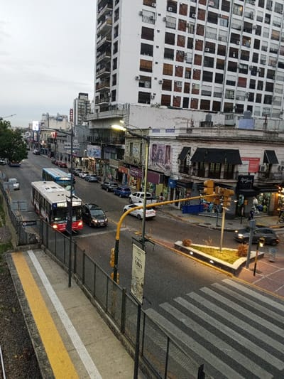 The image depicts an urban street scene in Ramos Mejía, Argentina, observed from an elevated perspective under an overcast sky, with streetlights illuminated. In the immediate foreground, a concrete platform features yellow and white tactile paving, separated from the roadway by a black metal fence.
A multi-lane street extends from the left to the right, intersecting with another street entering from the right. A red and white articulated bus, marked "38" and "73", is positioned on the left side of the main street, with its headlights illuminated. A dark SUV is adjacent to the bus, and a blue bus is partially visible behind it. Multiple vehicles, including parked and moving cars, are present throughout the street and at the intersection. A white sedan is positioned at the intersection, while a dark gray car is observed executing a right turn in the mid-right.
Buildings line the street, housing various commercial establishments at ground level, indicated by storefronts with visible signage such as "Fravega" and "Anjou Cosmetique." A prominent, tall, multi-story white building with numerous rectangular windows, many fitted with dark shutters or blinds, dominates the upper right background. Pedestrians are visible walking on the sidewalks and near the intersection. The intersection is equipped with yellow traffic light poles, one of which displays a vertical banner. A small, triangular planter containing sparse vegetation is situated in a median at the intersection. A marked zebra crossing is visible in the lower right foreground.